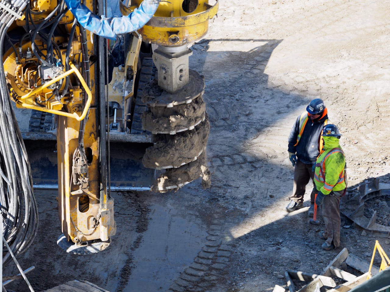 services-02 Two workers in protective gear at a construction site with heavy drilling machinery.